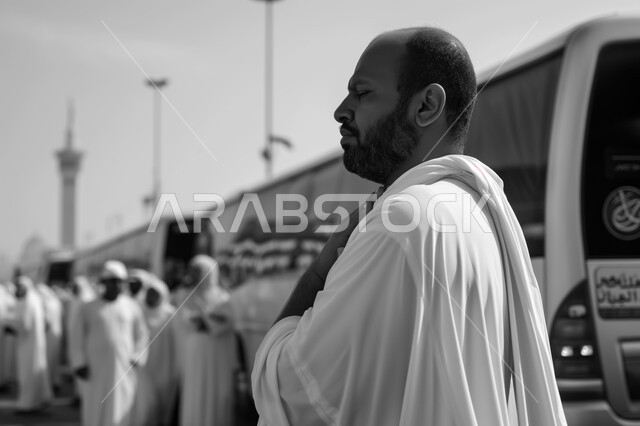 Visiting religious places, performing Hajj and Umrah rituals, sacred Islamic places and landmarks, worshiping and getting closer to God Almighty, a close-up picture from the side in black and white of an elderly Saudi Gulf Arab man wearing a white abaya standing at the bus stop of the Holy Mosque of Mecca in Mecca.