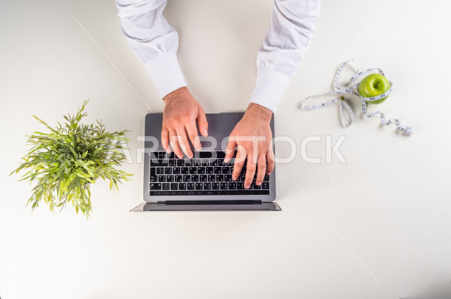 صورة مقربة من الأعلى , لأخصائي تغذية يعمل من خلال اللابتوب على مكتب أبيض , مهنة ووظيفة , بيئة عمل , خلفية بيضاء . 