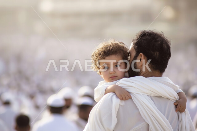Visiting religious places, worshiping and getting closer to God Almighty, performing Hajj and Umrah rituals, holy Islamic monuments, a close-up photo from the back of a veiled Saudi Arabian Gulf woman wearing a white abaya standing in the Holy Mosque of Mecca in Mecca holding her child in her arms