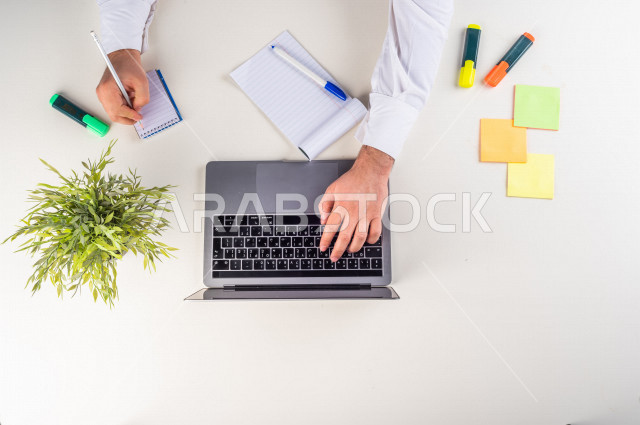 A close-up image from the top, of a Saudi Arab employee working on a laptop on a white desk, professions and jobs, a work environment, a white background.