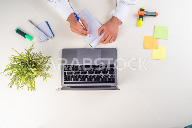 صورة مقربة من الأعلى , لموظف سعودي عربي يعمل من خلال اللابتوب على مكتب أبيض , مهن ووظيفة , بيئة عمل , خلفية بيضاء .