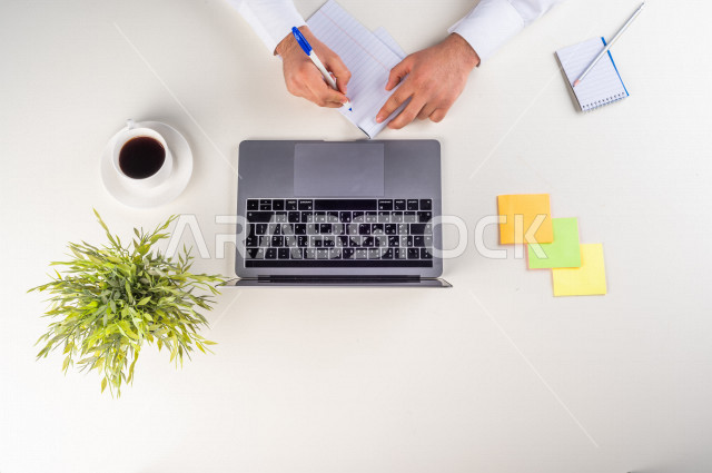 صورة مقربة من الأعلى , لموظف سعودي عربي يعمل من خلال اللابتوب على مكتب أبيض , مهن ووظيفة , بيئة عمل , خلفية بيضاء .