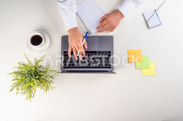 صورة مقربة من الأعلى , لموظف سعودي عربي يعمل من خلال اللابتوب على مكتب أبيض , مهن ووظيفة , بيئة عمل , خلفية بيضاء .