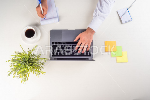 صورة مقربة من الأعلى , لموظف سعودي عربي يعمل من خلال اللابتوب على مكتب أبيض , مهن ووظيفة , بيئة عمل , خلفية بيضاء .