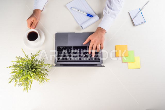 صورة مقربة من الأعلى , لموظف سعودي عربي يعمل من خلال اللابتوب على مكتب أبيض , مهن ووظيفة , بيئة عمل , خلفية بيضاء .