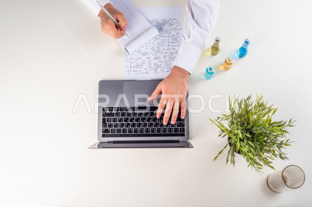 صورة مقربة من الأعلى , لأستاذ متخصص بالكيمياء يعمل من خلال اللابتوب على مكتب أبيض , بيئة عمل , خلفية بيضاء . 