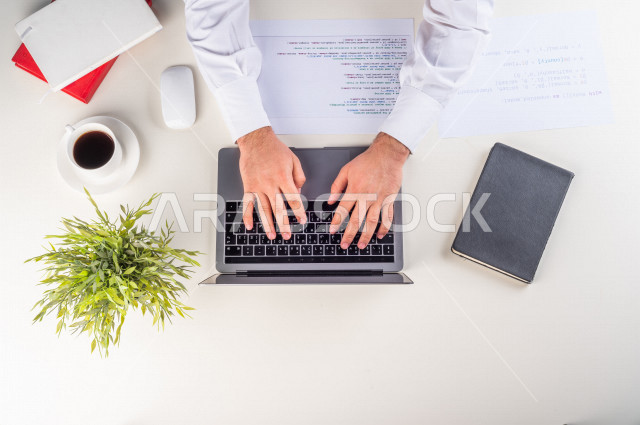 صورة مقربة من الأعلى , لموظف سعودي عربي يعمل من خلال اللابتوب على مكتب أبيض , مهن ووظيفة , بيئة عمل , خلفية بيضاء .