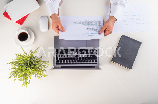 صورة مقربة من الأعلى , لموظف سعودي عربي يعمل من خلال اللابتوب على مكتب أبيض , مهن ووظيفة , بيئة عمل , خلفية بيضاء .