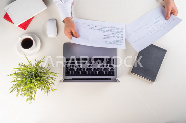 صورة مقربة من الأعلى , لموظف سعودي عربي يعمل من خلال اللابتوب على مكتب أبيض , مهن ووظيفة , بيئة عمل , خلفية بيضاء .