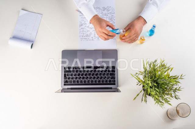 صورة مقربة من الأعلى , لأستاذ متخصص بالكيمياء يعمل من خلال اللابتوب على مكتب أبيض , بيئة عمل , خلفية بيضاء . 