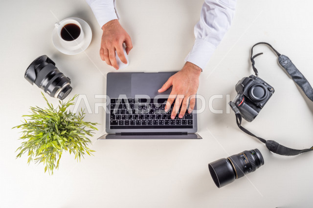  تصوير مقرب من الأعلى , لمصور عربي سعودي لديه خبره في التقاط الصور , مصور فوتوجرافيا يدخل الصور من خلال اللابتوب على مكتب أبيض , وظيفه ومهن , بيئة عمل , خلفية بيضاء .