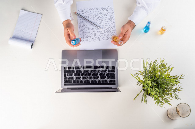 صورة مقربة من الأعلى , لأستاذ متخصص بالكيمياء يعمل من خلال اللابتوب على مكتب أبيض , بيئة عمل , خلفية بيضاء . 