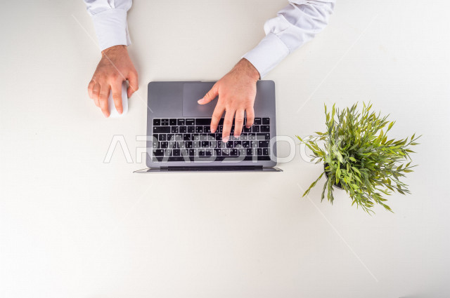 صورة مقربة من الأعلى , لموظف سعودي يعمل من خلال اللابتوب على مكتب أبيض , وظيفة ومهن , بيئة عمل , خلفية بيضاء .