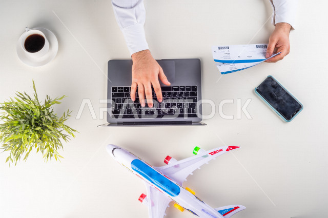 صورة مقربة من الأعلى , لرجل خليجي سعودي يضع معلومات تذاكر الرحلات الى اللابتوب في المطار , بيئة عمل , خلفية بيضاء .