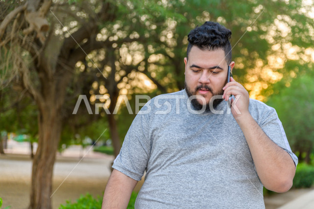 رجل شاب سعودي خليجي يمارس الرياضة الصباحية في الهواء الطلق ، و يقوم باجراء مكالمة صوتية عبر هاتفه المحمول  ،ممارسة رياضة المشي ، ممارسة الرياضة على طريق الممشى المخصص للمشي ، نشاط بدني  للمحافظة على اللياقة و الصحة الجيدة ، ممارسة التمارين الرياضية 