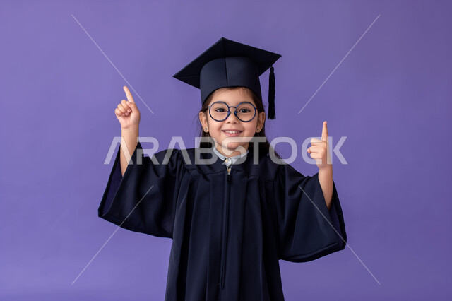 الترفع الى سنة دراسية جديدة، الانتهاء من الفصل الدراسي، النظر الى الكاميرا بايماءات الفخر والسعادة، الإحتفال بتحقيق النجاح، بورتريه مقرب لطالبة مدرسية عربية خليجية سعودية ترتدي ثوب و قبعة التخرج ترفع اصبعيها السبابة للأعلى، خلفية بنفسجي