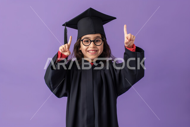 الإنتقال الى سنة دراسية جديدة، الانتهاء من الفصل الدراسي، النظر الى الكاميرا بايماءات الفخر والسعادة، الإحتفال بتحقيق النجاح، بورتريه مقرب لطالبة مدرسية عربية خليجية سعودية ترتدي ثوب و قبعة التخرج ترفع اصبعيها السبابة للأعلى، خلفية بنفسجي