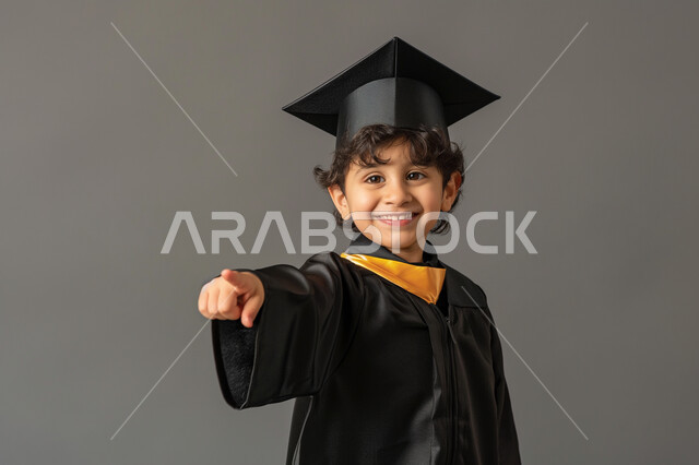 Gestures of pride and happiness to achieve success, the concept of education in the Kingdom of Saudi Arabia, celebrating graduation from the classroom, a portrait of a Saudi Gulf Arab school student wearing a graduation gown and cap, pointing with his hand to something, moving to a new school year, gray background