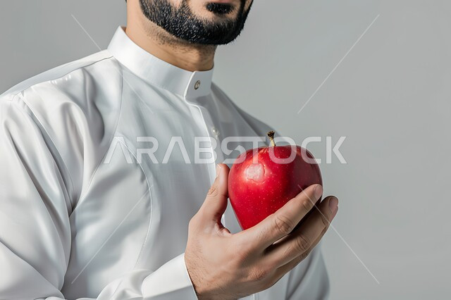 وجبات دايت صحية خفيفة، محاصيل زراعية سعودية ذات جودة عالية، تحسين مستويات الطاقة والتركيز خلال اليوم من خلال تناول الفواكه، بورتريه مقرب لرجل عربي سعودي خليجي يرتديالز يحمل بيده حبة من التفاح الاحمر، خلفيه رماديه