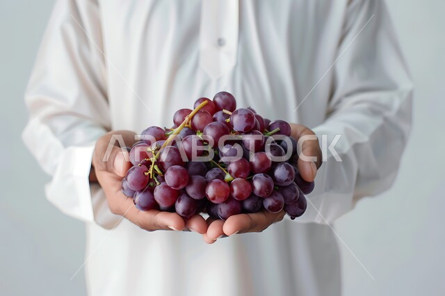 تحسين مستويات الطاقة والتركيز خلال اليوم من خلال تناول الفواكه، وجبات دايت صحية خفيفة، محاصيل زراعية سعودية ذات جودة عالية، بورتريه مقرب لرجل عربي سعودي خليجي يرتدي الزي التقليدي يحمل بيده مجموعة من حبات العنب، خلفيه رماديه