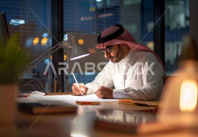 ايماءات الجديه والتركيز في العمل الليلي، مهن ووظائف ادارية مكتبية، رجل أعمال عربي خليجي سعودي يرتدي الزي التقليدي والشماغ يقوم بدراسة جدول الاعمال