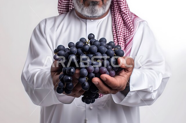 محاصيل زراعية سعودية ذات جودة عالية، تحسين مستويات الطاقة والتركيز خلال اليوم من خلال تناول الفواكه، وجبات دايت صحية خفيفة، بورتريه مقرب لرجل عربي سعودي خليجي يرتدي الزي التقليدي يحمل بيده مجموعة من حبات العنب، خلفيه رماديه