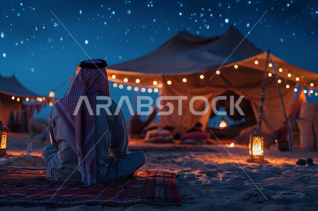 خيمة بدوية شعبية مضاءة بالفوانيس المعدنية وسط الصحراء، صورة مقربة من الخلف لرجل عربي خليجي سعودي يرتدي الشماغ والثوب التقليدي يجلس على الرمال الذهبية الناعمة في وقت الليل، كشتة و جلسة بر شبابية، التخييم في صحاري المملكة, مخيمات سياحية في المناطق البرية