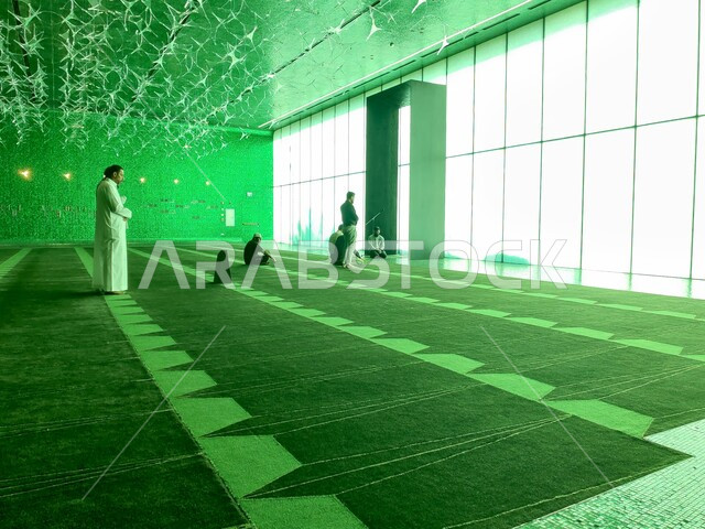 مجموعة رجال مسلمون عرب خليجييون سعودييون يؤدون فروض الصلاة داخل مصلى مسجد مركز الملك عبد العزيز الثقافي العالمي المضاء باللون الأخضر، مركز إثراء في مدينة الظهران في المملكة العربية السعودية، أماكن دينية إسلامية العبادة والتقرب إلى الله