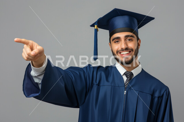 مفهوم الاجتهاد والتفوق، الاحتفال بإنهاء الدراسة الجامعية، بورتريه مقرب لطالب عربي سعودي خليجي يرتدي العباءه وقبعة التخرج ينظر إلى الكاميرا يشير بإصبع السبابة الى شيء ما بإيماءات السرور, الشعور بالفرح والذهول, خلفية رمادية