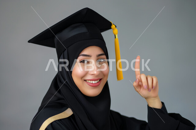 الشعور بالفرح والسرور, الاحتفال بإنهاء الدراسة الجامعية، مفهوم الاجتهاد والتفوق، بورتريه مقرب لطالبة عربية سعودية خليجية ترتدي العباءه و قبعة التخرج تنظر إلى الكاميرا تشير بإصبع السبابة الى شيء ما بإيماءات السعادة والثقة بالنفس, خلفية رمادية
