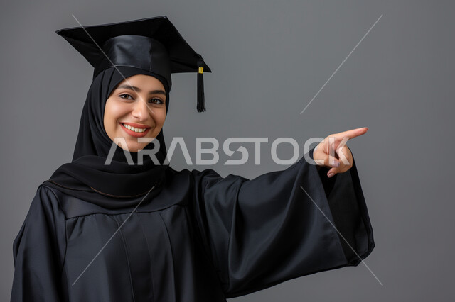 الشعور بالفرح والسرور, الاحتفال بإنهاء الدراسة الجامعية، مفهوم الاجتهاد والتفوق، بورتريه مقرب لطالبة عربية سعودية خليجية ترتدي العباءه و قبعة التخرج تنظر إلى الكاميرا تشير بإصبع السبابة الى شيء ما بإيماءات السعادة والثقة بالنفس, خلفية رمادية