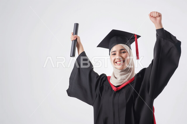 النجاح والاجتهاد والتفوق، الشعور بالفرح والسرور, الاحتفال بإنهاء الدراسة الجامعية، بورتريه مقرب لطالبة عربية سعودية خليجية ترتدي العباءه و قبعة التخرج تنظر إلى الكاميرا ترفع يديها للاعلى تمسك الشهادة بإيماءات السعادة, خلفية رمادية