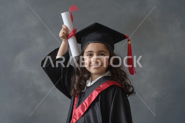 النجاح والاجتهاد والتفوق، الشعور بالفرح والسرور, بورتريه مقرب لطالبة عربية سعودية خليجية ترتدي العباءه و قبعة التخرج تنظر إلى الكاميرا ترفع يديها للاعلى تمسك الشهادة بإيماءات السعادة, خلفية رمادية