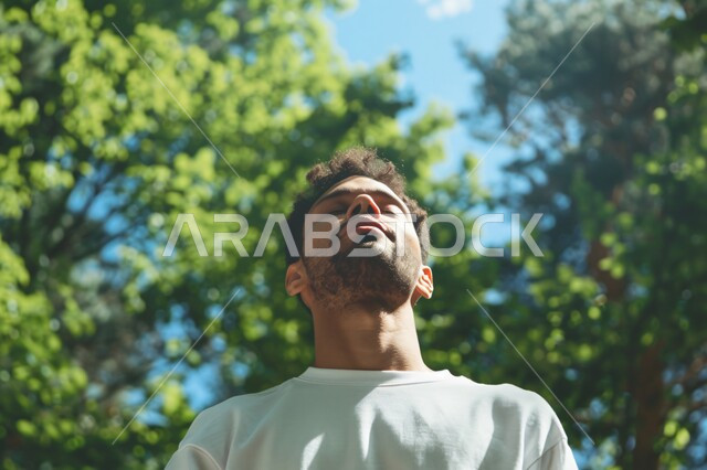 الاستمتاع في قضاء الأوقات في العطلة الصيفية، الشعور بالراحة والتأمل والاستجمام، المشي و التنزه في الحدائق العامة لاستنشاق الهواء الطلق ، صورة مقربة لشاب عربي خليجي سعودي مبتسم يغمض عينيه بايماءات التخيل، الطبيعة الخضراء في المملكة العربية السعودية