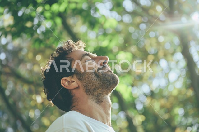 الشعور بالراحة والتأمل والاستجمام، المشي و التنزه في الحدائق العامة لاستنشاق الهواء الطلق ، صورة مقربة لشاب عربي خليجي سعودي مبتسم يغمض عينيه بايماءات التخيل، قضاء أوقات ممتعة في العطلة الصيفية، الطبيعة الخضراء في المملكة العربية السعودية