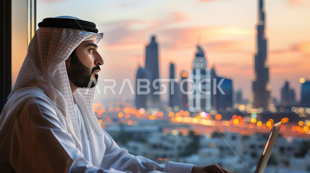 إنجاز الأعمال باستخدام الأجهزة التقنية، بيئة العمل المريحة في الشركات السعودية، تطوير الخطط وحل المشكلات لتحقيق أهداف الشركة، موظف عربي خليجي سعودي يقوم بإدارة شؤون العمل وتنظيمه عبر الحاسوب المحمول بجانب نافذة مطلة على المدينة