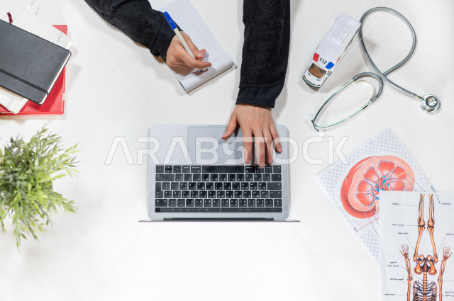  تصوير مقرب من الأعلى , لطبيبة عربية سعودية لديها خبره في طب الباطنية , طبيبة باطنية تعمل من خلال اللابتوب على مكتب أبيض , وظيفه ومهن , بيئة عمل , خلفية بيضاء .