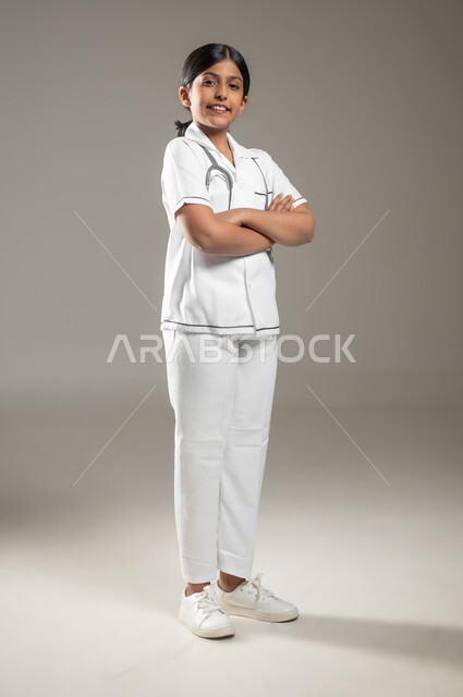 الوقوف باستقامة والنظر الى الكاميرا بإيماءات أيدي متقاطعة، الطموح في تحقيق الآمال، الشغف بالحصول على مهنة الطب منذ الصغر، بورتريه لفتاة عربية خليجية سعودية مبتسمة ترتدي المعطف الأبيض وسماعة الطبيب، الجسم بالطول الكامل، خلفية رمادية، متحوى حساس، استخدام حساس