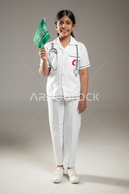 يوم العلم 11 مارس، التعبير عن الولاء للدولة في اليوم الوطني السعودي 23 سبتمبر, بورتريه مقرب لفتاة عربية خليجية سعودية مبتسمة ترتدي المعطف الطبي الأبيض تحلم أن تصبح طبيبة طوارئ تمسك علم المملكة, الجسم بالطول الكامل، خلفية رمادية، محتوى حساس، استخدام حساس