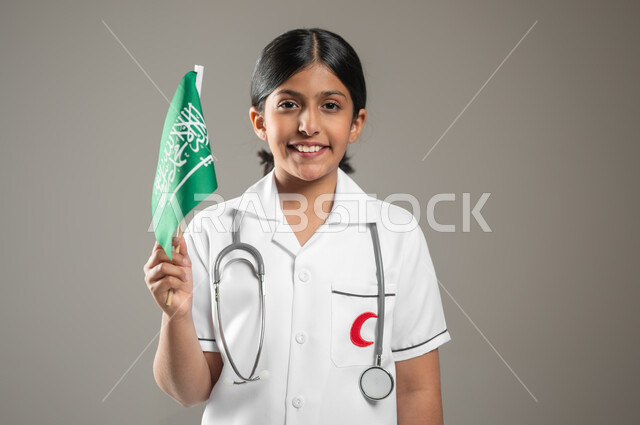 التعبير عن الولاء للدولة في اليوم الوطني السعودي 23 سبتمبر,  بورتريه مقرب لفتاة عربية خليجية سعودية مبتسمة ترتدي المعطف الطبي الأبيض وسماعة الطبيب تحلم أن تصبح طبيبة طوارئ وإسعاف تمسك علم المملكة, يوم العلم 11 مارس, خلفية رمادية، محتوى حساس، استخدام حساس