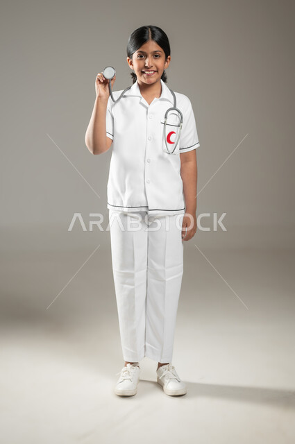 النظر إلى الكاميرا بإيماءات الشغف، استخدام سماعة نبضات القلب، بورتريه  لفتاة عربية خليجية سعودية مبتسمة ترتدي زي الطبيب تحلم أن تصبح طبيبة محترفة، اختيار المهنة المستقبلية منذ الصغر،  الجسم بالطول الكامل، خلفية رمادية، استخدام حساس، محتوى حساس