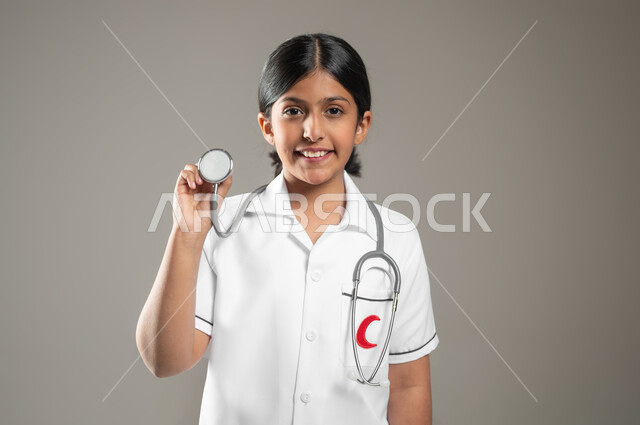 طموحات وأهداف طفولية، استخدام سماعة نبضات القلب، النظر إلى الكاميرا بإيماءات الشغف، بورتريه مقرب لفتاة عربية خليجية سعودية مبتسمة ترتدي زي الطبيب تحلم أن تصبح طبيبة محترفة، اختيار المهنة المستقبلية منذ الصغر، خلفية رمادية، استخدام حساس، محتوى حساس