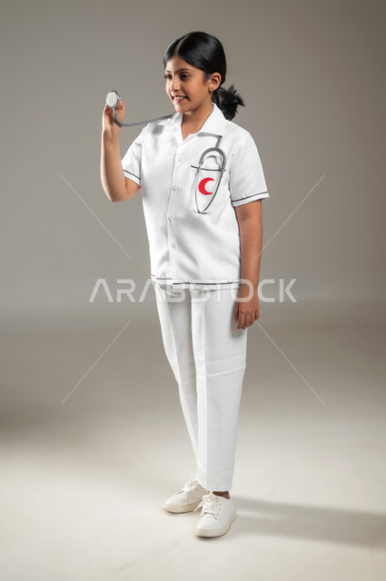 اختيار المهنة المستقبلية منذ الصغر، النظر إلى سماعة نبضات القلب بإيماءات الشغف بتحقيق حلم الطفولة، تقديم الخدمات للمرضى، بورتريه لفتاة عربية خليجية سعودية مبتسمة ترتدي زي الطبيب تحلم أن تصبح طبيبة، الجسم بالطول الكامل، خلفية رمادية، استخدام حساس، محتوى حساس