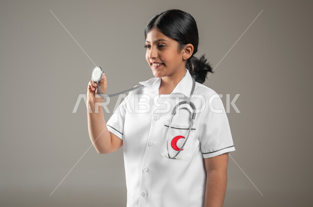 النظر إلى سماعة نبضات القلب بإيماءات الشغف  بتحقيق حلم الطفولة، تقديم الخدمات للمرضى، بورتريه مقرب لفتاة عربية خليجية سعودية مبتسمة ترتدي زي الطبيب تحلم أن تصبح طبيبة، اختيار المهنة المستقبلية منذ الصغر، خلفية رمادية، استخدام حساس، محتوى حساس