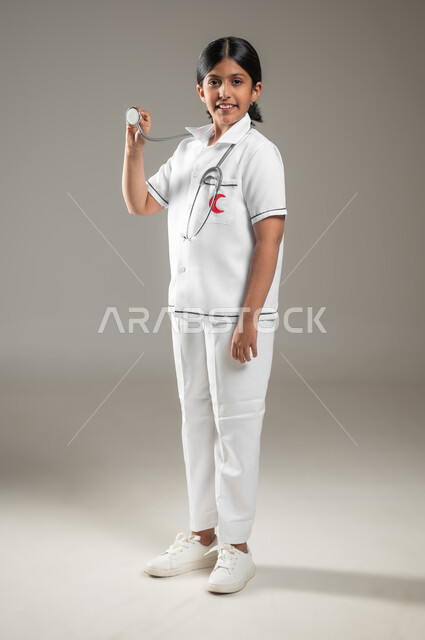 تقديم الخدمات للمرضى، استخدام سماعة نبضات القلب، النظر إلى الكاميرا بإيماءات الشغف، بورتريه لفتاة عربية خليجية سعودية مبتسمة ترتدي زي الطبيب تحلم أن تصبح طبيبة، اختيار المهنة المستقبلية منذ الصغر، الجسم بالطول الكامل، خلفية رمادية، استخدام حساس، محتوى حساس