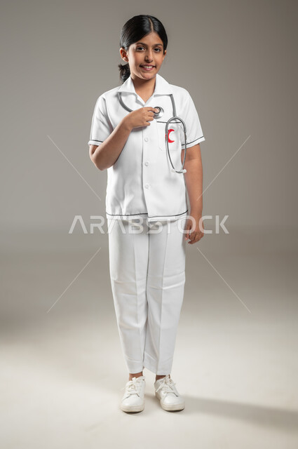 طموحات وأهداف طفولية، استخدام سماعة نبضات القلب، النظر إلى الكاميرا بإيماءات الشغف، بورتريه لفتاة عربية خليجية سعودية مبتسمة ترتدي زي الطبيب تحلم أن تصبح طبيبة، اختيار المهنة المستقبلية منذ الصغر، الجسم بالطول الكامل، خلفية رمادية، استخدام حساس، محتوى حساس