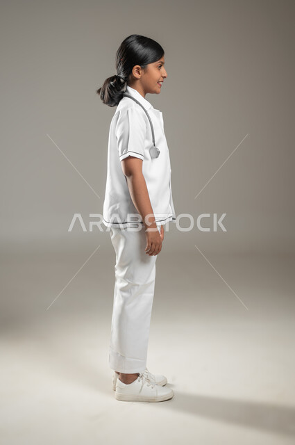 تقديم الخدمات الطبية والاستعداد لحالات الطوارئ, النظر الى مكان ما بايماءات البشاشه، بورتريه من الجانب لفتاة عربية خليجية سعودية ترتدي اللباس الطبي مع السماعة الطبية، الجسم بالطول الكامل، خلفية رمادية، محتوى حساس، استخدام حساس