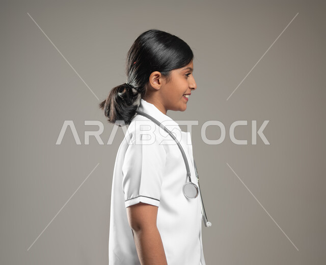 النظر الى مكان ما بايماءات البشاشه، الاهتمام بتقديم الخدمات الطبية والاستعداد لحالات الطوارئ, بورتريه مقرب من الجانب لفتاة عربية خليجية سعودية ترتدي اللباس الطبي مع السماعة الطبية، خلفية رمادية، محتوى حساس، استخدام حساس