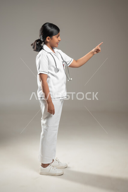 تقديم الخدمات الطبية والاستعداد لحالات الطوارئ, النظر إلى الكاميرا، بورتريه من الجانب لفتاة عربية خليجية سعودية ترتدي اللباس الطبي وتشير باصبعها الى مكان ما، الجسم بالطول الكامل، خلفية رمادية، محتوى حساس، استخدام حساس