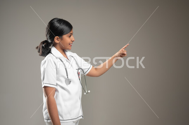 الاهتمام بتقديم الخدمات الطبية والاستعداد لحالات الطوارئ, النظر إلى الكاميرا، بورتريه مقرب من الجانب لفتاة عربية خليجية سعودية ترتدي اللباس الطبي  وتشير باصبعها الى مكان ما، خلفية رمادية، محتوى حساس، استخدام حساس